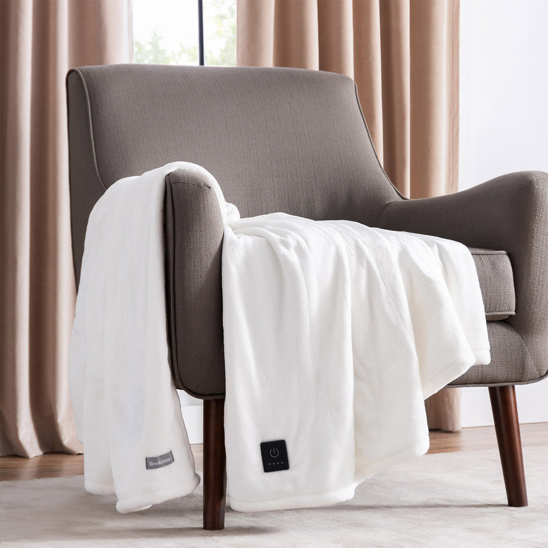 Ivory oversized heated throw draped over a modern gray armchair, showing the built-in control button.