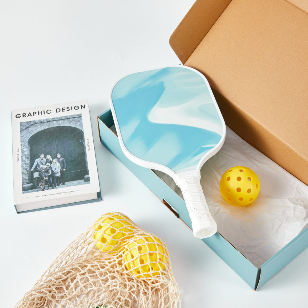 Blue paddle with a yellow ball on a white surface next to a box and photo album.