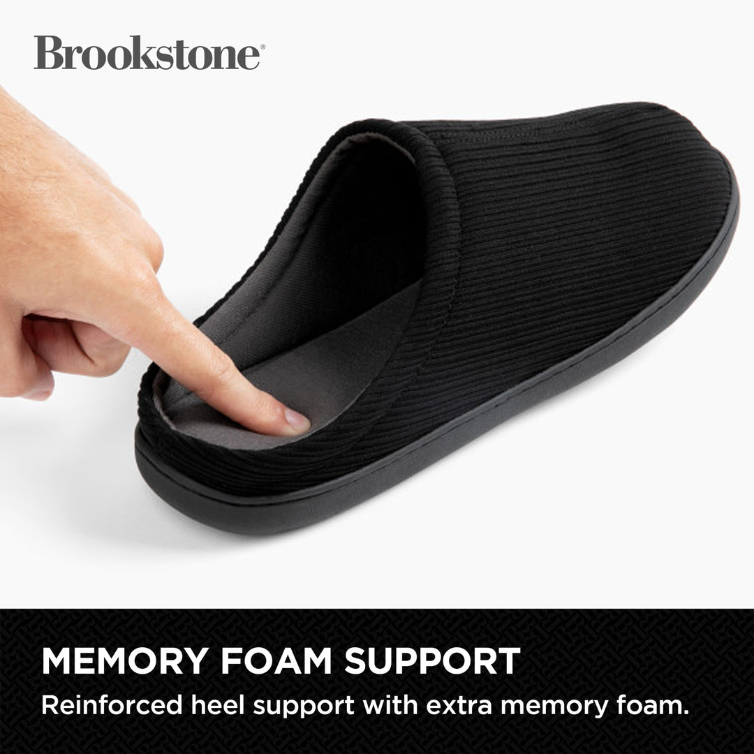 Black slipper with memory foam support being touched by a hand, Brookstone brand visible.