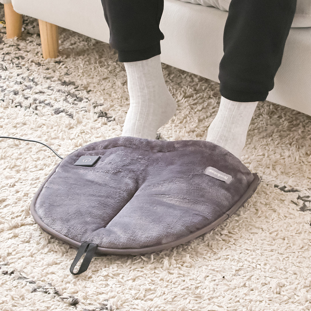 Gray foot warmer pad on a carpeted floor with a person wearing socks.