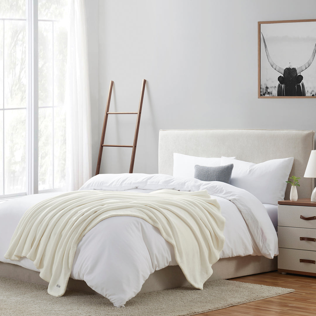 Neatly made bed with white bedding and a beige throw blanket in a bright bedroom.