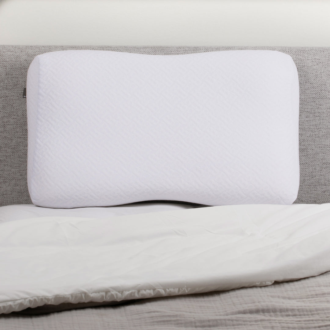White contoured memory foam pillow resting against a gray headboard on a neatly made bed.