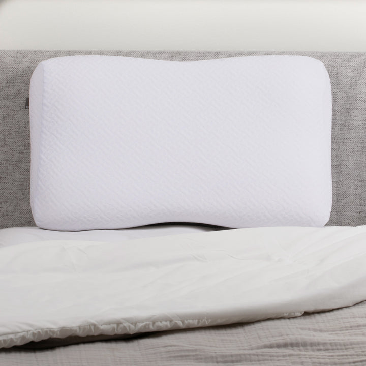 White contoured memory foam pillow resting against a gray headboard on a neatly made bed.
