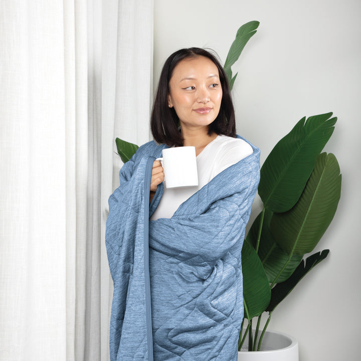 Woman wrapped in a blue blanket holding a white mug, standing indoors with plants in the background.