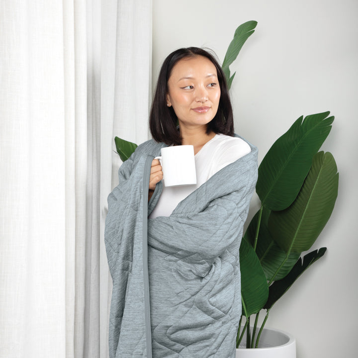 Woman wrapped in a gray blanket holding a white mug, standing next to a plant indoors.
