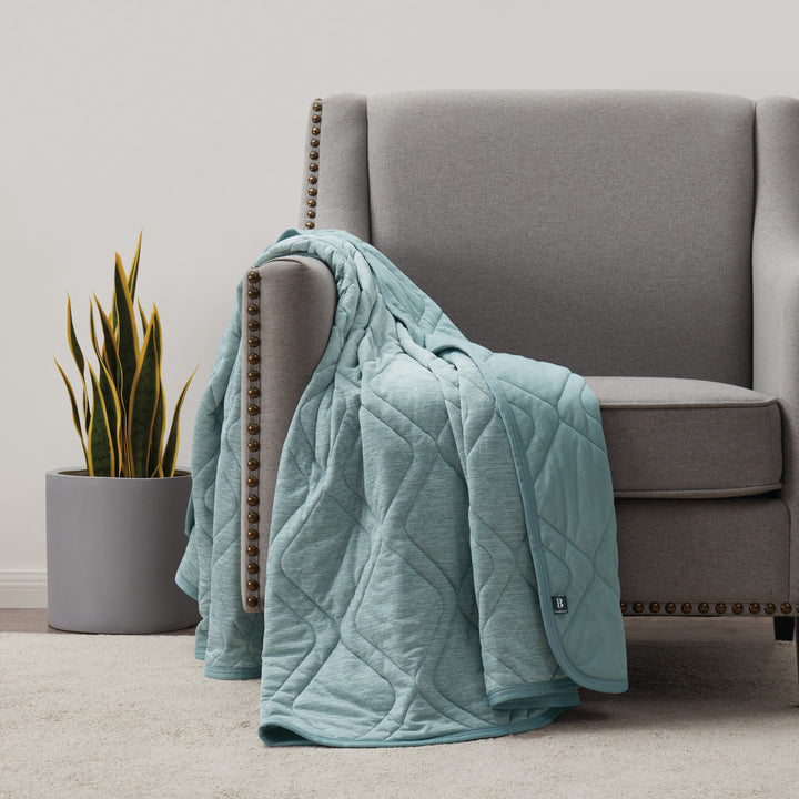 Light blue quilted blanket draped over a gray armchair in a room with a plant.