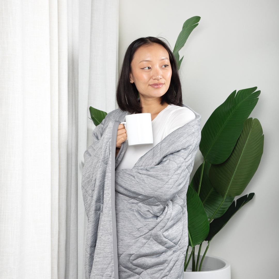 Woman wrapped in a gray blanket holding a white mug, standing next to a plant indoors.