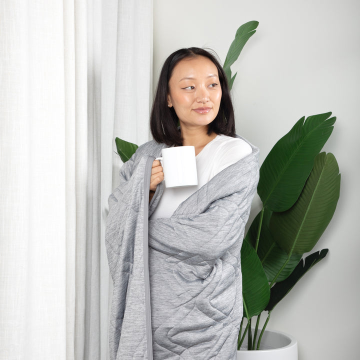 Woman wrapped in a gray blanket holding a white mug, standing next to a plant indoors.