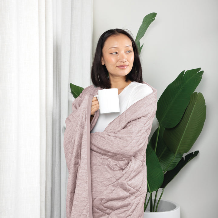 Woman wrapped in a pink blanket holding a white mug, standing next to a plant indoors.