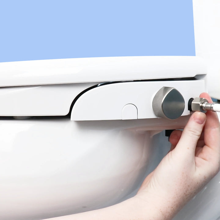 Close-up of a TUSHY Oasis platinum control knob  with a hand attaching the water hose, on a white background