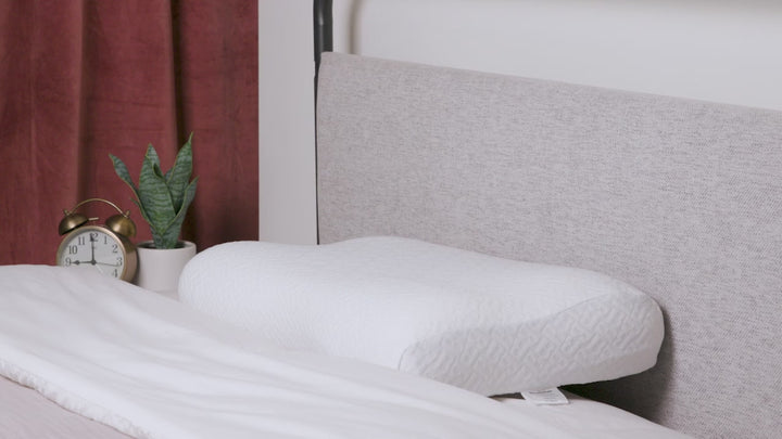 Video showing a contoured white memory foam pillow on a bed beside a gold alarm clock and a potted plant, with a light gray headboard and rust-colored curtains in the background.